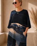 May include: A loose-fitting, short-sleeved dress featuring a dark navy blue base with a prominent graphic print of a whale. The design incorporates swirling, abstract lines in shades of light blue, cream, and white, creating a fluid, artistic pattern.