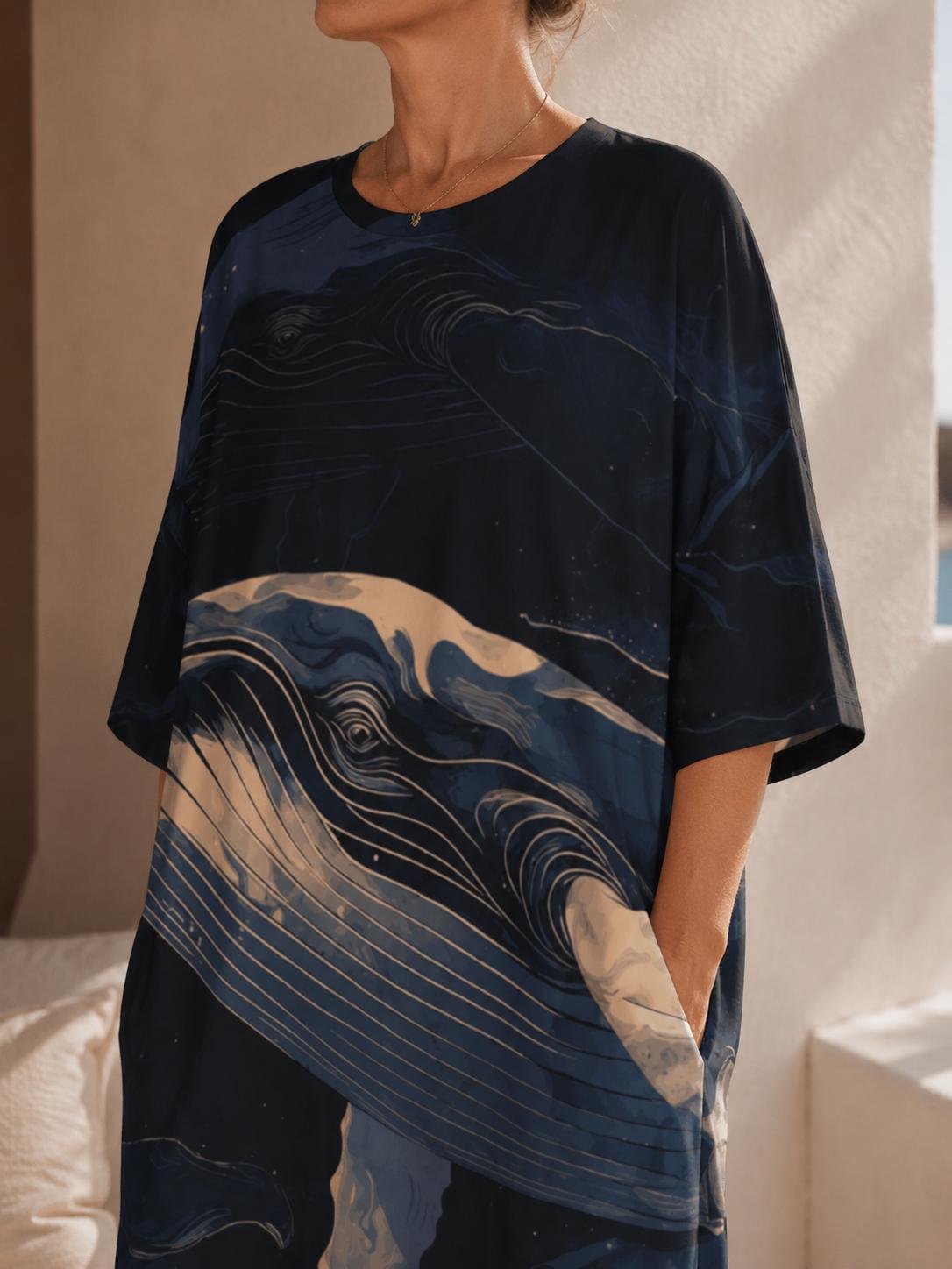 May include: A loose-fitting, short-sleeved tunic featuring a dark navy blue background with a large, stylized graphic print of a whale swimming through ocean waves in shades of light blue, cream, and white, with a round neckline and side pockets.